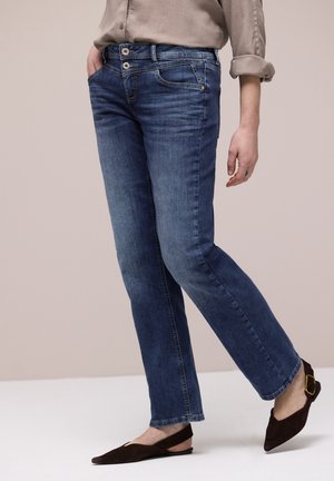 Person trägt blaue Jeans mit geradem Bein, Doppelknopfverschluss an der Taille und dunkelbraunen Slingback-Ballerinas, steht vor einem einfarbigen Hintergrund.