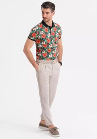 Blommig polo-shirt med gröna, röda och vita mönster, svart krage. Byxor är ljusbeige, med bälte. Skor är bruna i mocka.