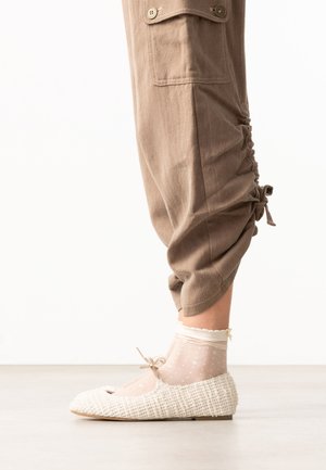 Lower leg wearing beige ruched pants, sheer white polka dot ankle socks, and white textured ballet flats with small bow.