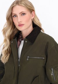 Olivgrüne Bomberjacke mit einem Cordkragen, Reißverschlusstaschen und einer glatten, matten Textur. Träger hat einen Schal unter dem Kragen.