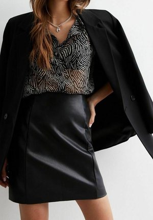 Nainen pukeutuneena mustaan bleiseriin, jossa on see-through seepra-kuosinen paita ja musta nahkainen hame, kaulassaan sydämenmuotoinen riipus.