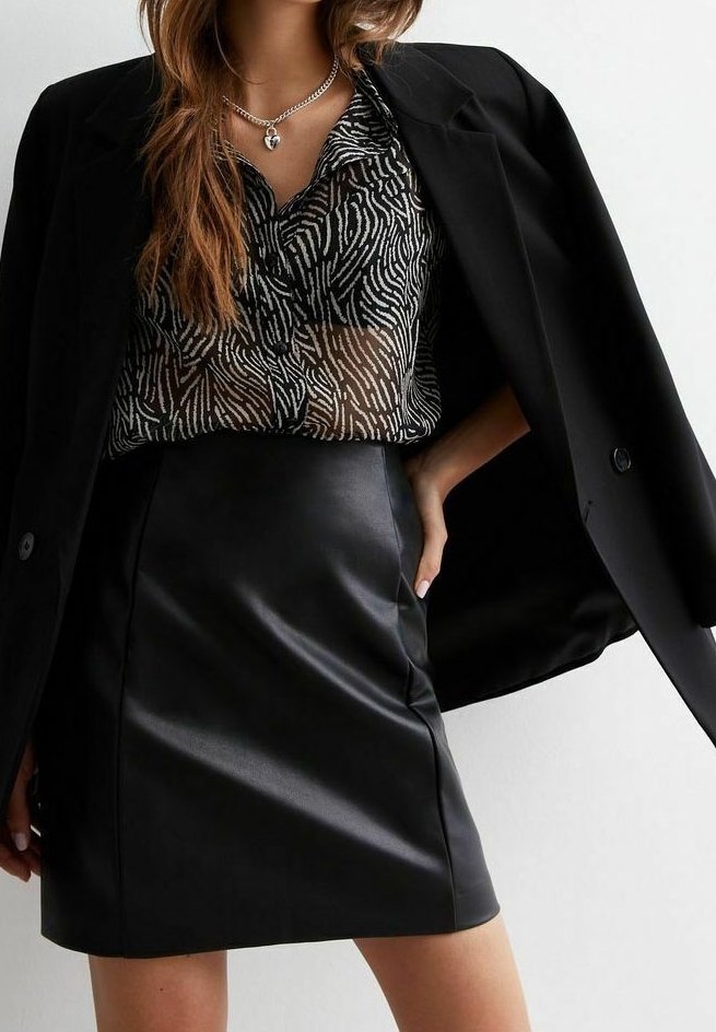 Femme portant un blazer noir sur un chemisier transparent à imprimé zèbre et une jupe en cuir noire, avec un collier pendentif en forme de cœur.
