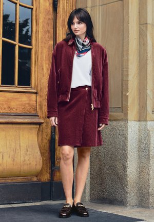 Una mujer con chaqueta y falda de color burdeos, un pañuelo estampado y mocasines negros está de pie junto a una puerta de madera.