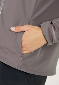 Grå letvægtsjakke med en lynlåslomme, der har en glat overflade og elastiske manchetter. Hånden er placeret inde i lommen.