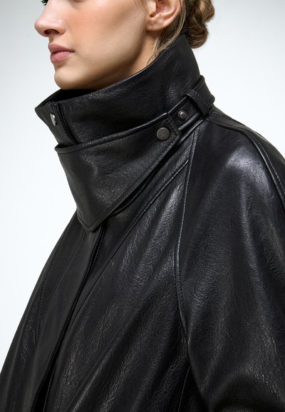 Mujer con chaqueta de cuero negra de cuello alto con detalles de botones a presión, de perfil contra un fondo liso.