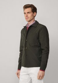 Ciemnozielona kurtka z kołnierzykiem, zapinana na guziki, z dwiema przednimi kieszeniami, wykonana z gładkiego, lekkiego materiału.