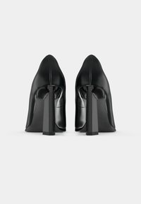 Scarpe nere in pelle con tacco alto e punta affilata, dotate di un ampio tacco quadrato e una superficie liscia e lucida. Dettaglio delle cuciture lungo il retro.