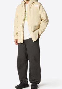 Mężczyzna ubrany w beżową kurtkę North Face na biały t-shirt z grafiką, luźne czarne spodnie oraz czarne buty na slip-on, stoi z rękami w kieszeniach.