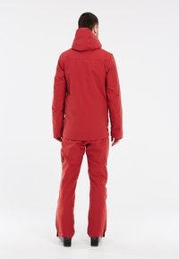 Rote wasserdichte Jacke mit verstellbarer Kapuze, kombiniert mit passenden roten Hosen. Die Hosen haben seitliche Reißverschlüsse. Glatte Textur, entworfen für den Außenbereich.