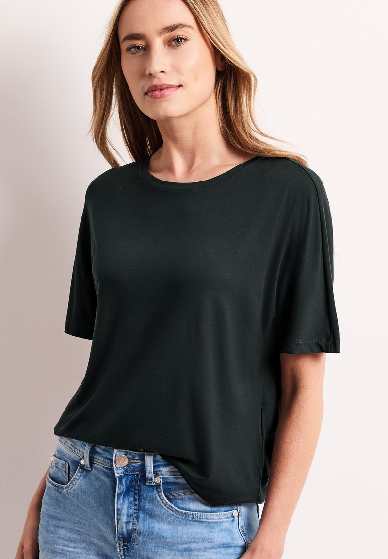 Femme aux cheveux blonds portant un t-shirt noir uni à manches courtes et un jean bleu clair, debout devant un fond neutre.