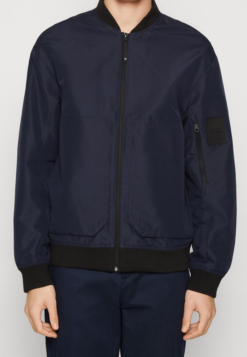 Blouson aviateur bleu marine en tissu lisse avec fermeture éclair frontale, poignets et ourlet côtelés. Comprend deux poches latérales et une poche zippée sur le bras.