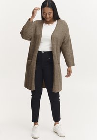 Brown knitted cardigan with textured sleeves, open front, and patch pockets, worn over a white lace-trimmed top and black pants.