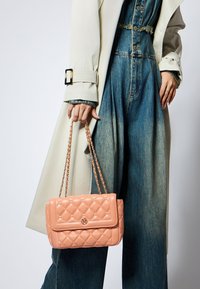 Różowa torebka pikowana z klapką, łańcuszkowym paskiem i dekoracyjnymi ćwiekami. Torebka ma emblemat logo z przodu, charakteryzujący się gładkim wykończeniem.