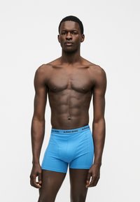 Λιγνός σκουρόχρωμος άνδρας στέκεται μπροστά σε απλό φόντο, φορώντας φωτεινά μπλε μποξέρ με λουρί με το λογότυπο "BJÖRN BORG".