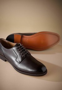 Zapatos de vestir de cuero negro con un acabado suave, punta redonda y diseño de cordones. Presentan una suela de cuero marrón en contraste con detalles de costura.