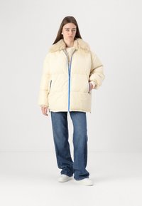 Light cream puffer jacket with a faux fur collar, blue zipper accent, side pockets, and a relaxed fit, paired with dark blue jeans.