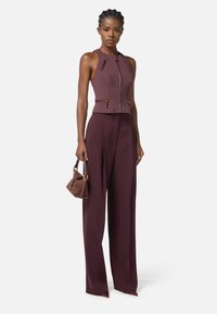 Top bordeaux aderente con scollatura alta e chiusura con zip, abbinato a pantaloni a gamba larga in vita alta dello stesso colore, mentre tiene una piccola borsa marrone.