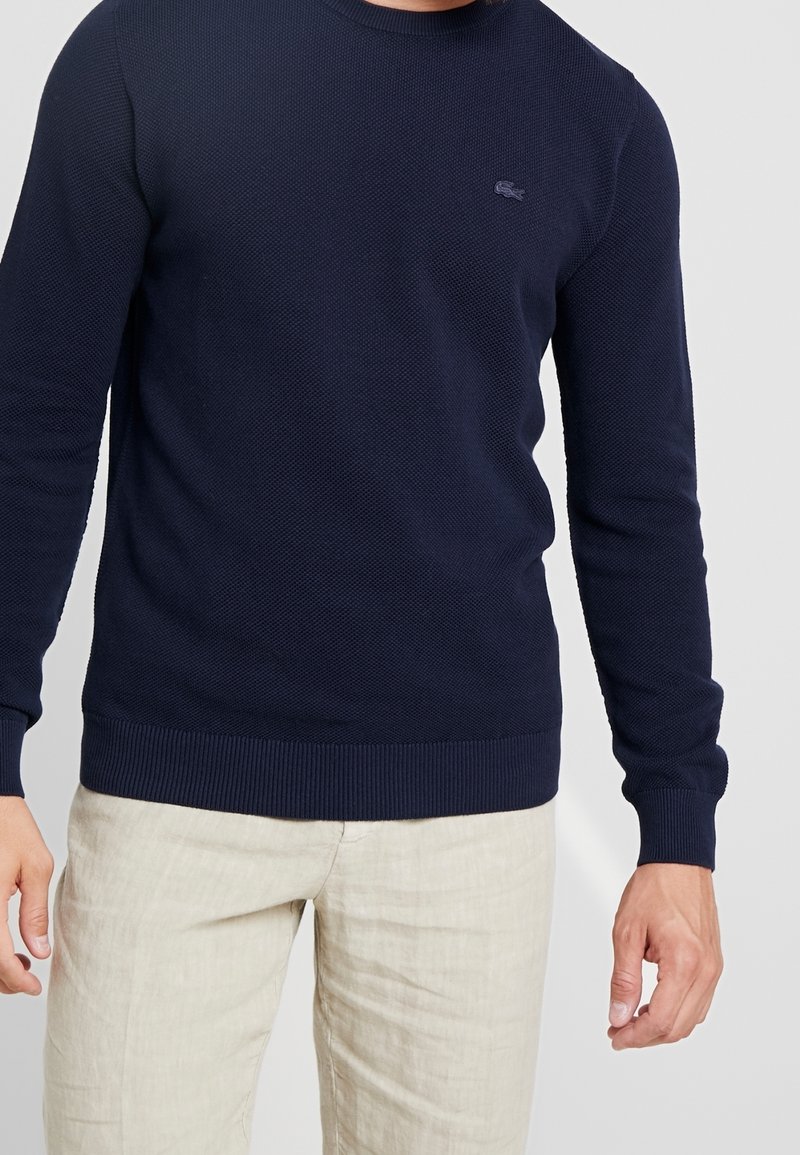 Homme portant un pull bleu marine texturé à manches longues avec un petit logo brodé sur la poitrine et un pantalon beige clair.