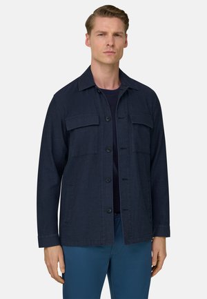 Mies, joka pukeutunut tummansiniseen napitettuun takkiin, mustaan paitaan ja sinisiin housuihin, seisoo yksinkertaista vaaleaa taustaa vasten.