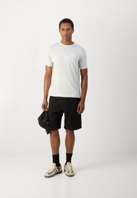 Lichtgrijs T-shirt met korte mouwen, zwarte shorts, groene en beige sneakers, houdt een zwarte pet vast. Eenvoudig ontwerp, casual pasvorm, minimale details.