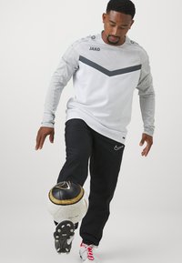 Witte sporttop met lange mouwen en grijze accenten, gecombineerd met zwarte broek. Speler balanceert een zwart en gouden voetbalt op een textuur oppervlak.