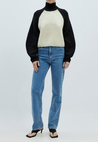 Gebreide coltrui met zwarte mouwen en een crèmekleurig lijf, gecombineerd met blauwe rechtlijnige jeans en zwarte hoge sandalen.