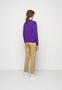 Person med lockigt hår, iförd en lila sweatshirt, beige byxor, vita strumpor och vita sneakers, stående vänd bort mot en grå bakgrund.