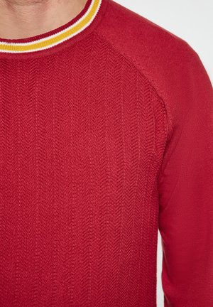 Roter Strickpullover mit einem strukturierten Zickzackmuster, ausgestattet mit einem gerippten Rundhalsausschnitt, der durch weiße und gelbe Streifen akzentuiert wird.