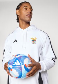 Felpa bianca con dettagli neri e logo Adidas, con emblema del Benfica. Il modello tiene in mano un pallone da calcio decorato con un motivo blu e rosso.