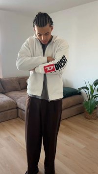 Young man with braided hair wearing a white Diesel hoodie and dark pants standing with arms crossed in a living room.