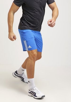 Mann trägt schwarzes T-Shirt, blaue Adidas-Shorts, weiße Adidas-Socken und weiße Adidas-Sneaker, geht auf einfarbigem Hintergrund.