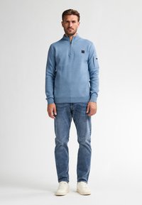 Pulover albastru deschis cu fermoar pe un sfert, manșete cu striații și un mic patch cu logo negru. Purtați-l cu blugi albaștri deschiși și adidași albi.