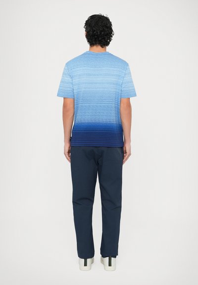 Zila gradienta t-krekls ar horizontālām svītrām, kas pāriet no gaiši zilas uz tumši zilu, kopā ar tumši ziliem biksēm un baltām kurpēm.