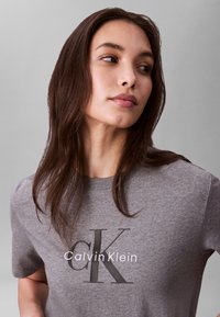 Fiatal nő hosszú barna hajjal, szürke Calvin Klein pólót visel, oldalra néz egy egyszínű háttér előtt.