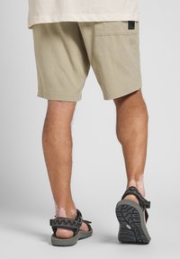 Beige shorts med et enkelt design, med en baklomme og en liten svart logo-patch. Brukt med mørke sandaler som viser teksturerte stropper.
