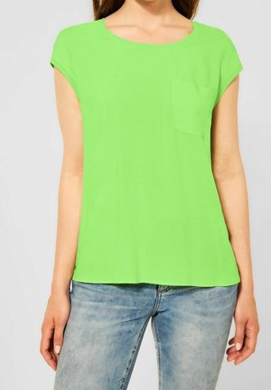 Frau trägt ein leuchtend grünes Kurzarmoberteil mit einer kleinen Brusttasche und hellblauen, ausgewaschenen Jeans.