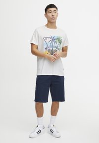 Lys grå t-skjorte med et palmegrafikk, kombinert med marineblå shorts. Modellen bruker hvite sokker og svartstripete hvite sneakers.