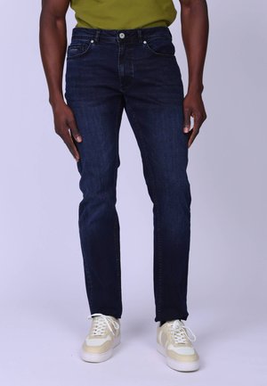 Homme portant un jean slim bleu foncé, une chemise verte et des baskets beiges, debout devant un fond clair uni.