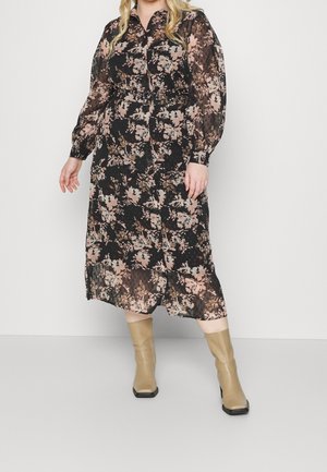 Frau trägt ein schwarzes, durchsichtiges Midikleid mit pinkem Blumenmuster und langen Ärmeln, kombiniert mit beigen Stiefeletten, stehend vor weißem Hintergrund.