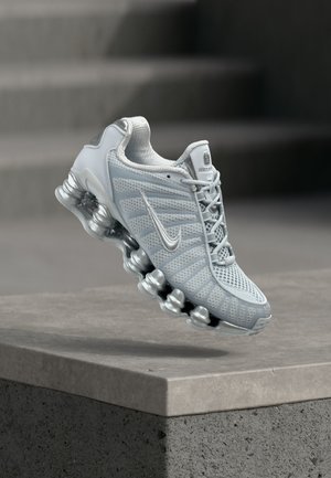 Chaussure de course Nike gris clair avec une semelle amortissante en forme de ressort flottant au-dessus d'un rebord en béton, arrière-plan d'escalier flou.