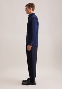 Marineblauwe kraagloze blouse met lange mouwen, in combinatie met donkere broeken en zwarte schoenen. Glad materiaal, tailored fit, minimale details.