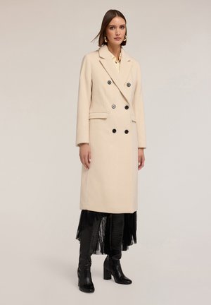 Cappotto beige in lana doppiopetto con bottoni neri, due tasche laterali, maniche lunghe e rever strutturati, abbinato a stivali in pelle neri.