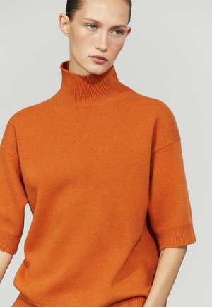Kvinde iført en orange turtleneck-trøje med korte ærmer, der kigger let til siden mod en ensfarvet lys baggrund.