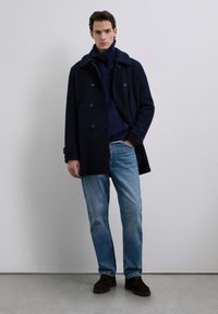 Mørkeblå uldfrakke med pelskrave, lagd over en mørk striktrøje, parret med lyseblå jeans og brune ruskindsko. Minimalt design.