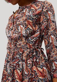Abito a maniche lunghe con motivo paisley in tonalità di rosso, blu navy e crema. Presenta un collo con bottoni e una vita regolabile con lacci. Tessuto dalla texture liscia.