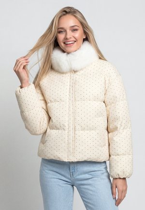 Lächelnde Frau mit langen blonden Haaren trägt eine cremefarbene Steppjacke mit weißem Kunstpelzkragen und hellblaue Jeans.