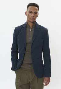 Blazer in cotone blu con due bottoni frontali, una tasca sul petto e pantaloni a pieghe verde oliva a contrasto indossati sopra un maglione in maglia marrone.