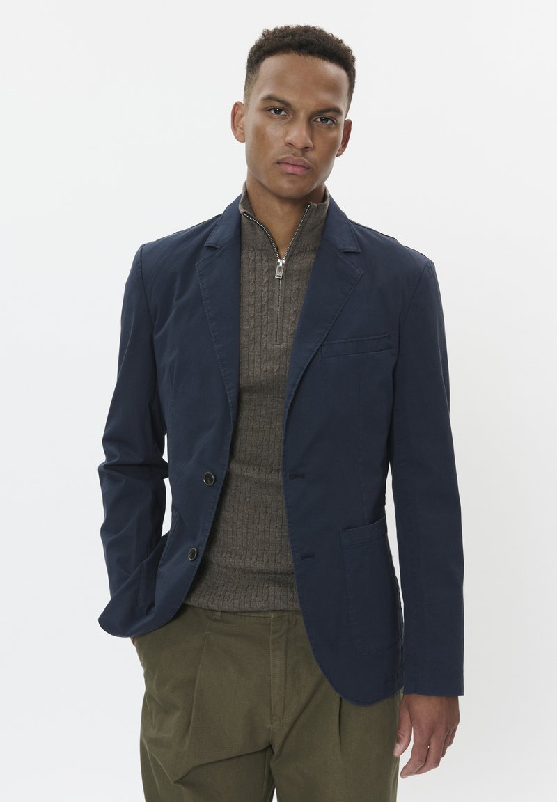 Blazer in cotone blu con due bottoni frontali, una tasca sul petto e pantaloni a pieghe verde oliva a contrasto indossati sopra un maglione in maglia marrone.