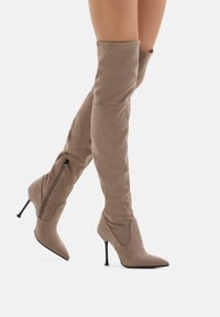 Stivali alti in suede taupe con punta affusolata, cerniere laterali e sottili tacchi a stiletto neri indossati su gambe incrociate.