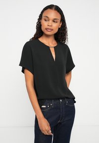 Woman with curly hair wearing a black short-sleeve blouse with a metal clasp at the neckline and dark blue jeans, standing against a white background.